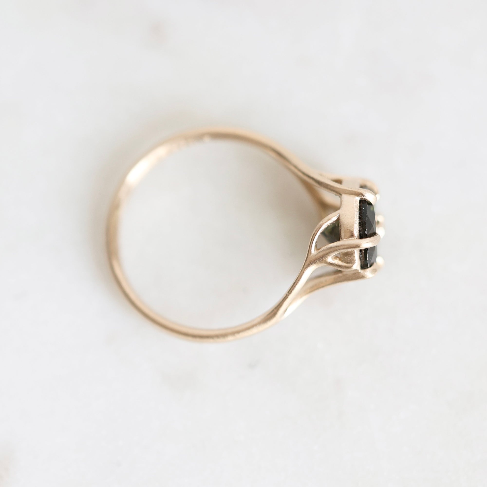 A gold ring with a small black rectangular gemstone in a prong setting, photographed from above on a light off-white background.