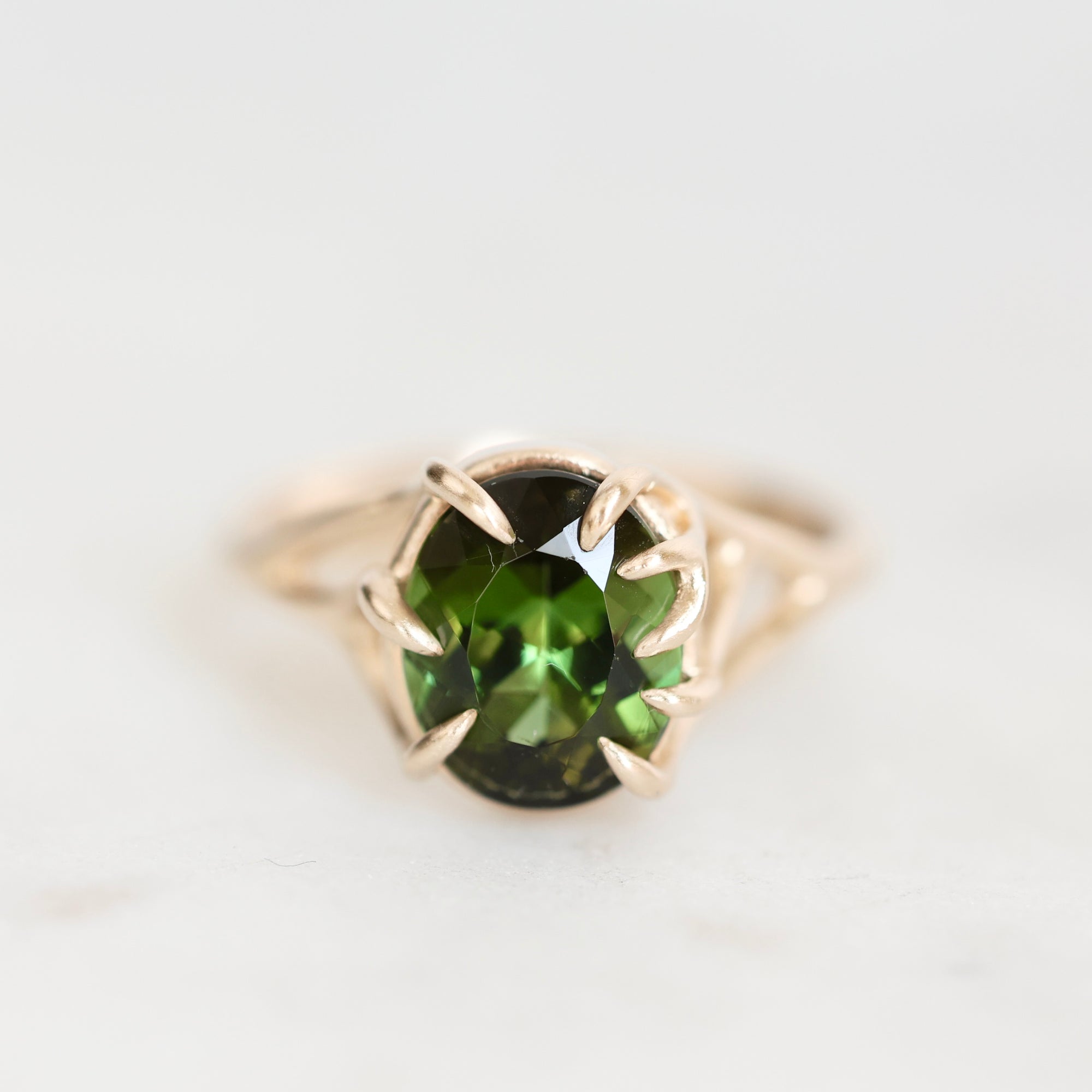 An oval-cut green gemstone ring set in a gold prong setting, shown on a light, neutral background.