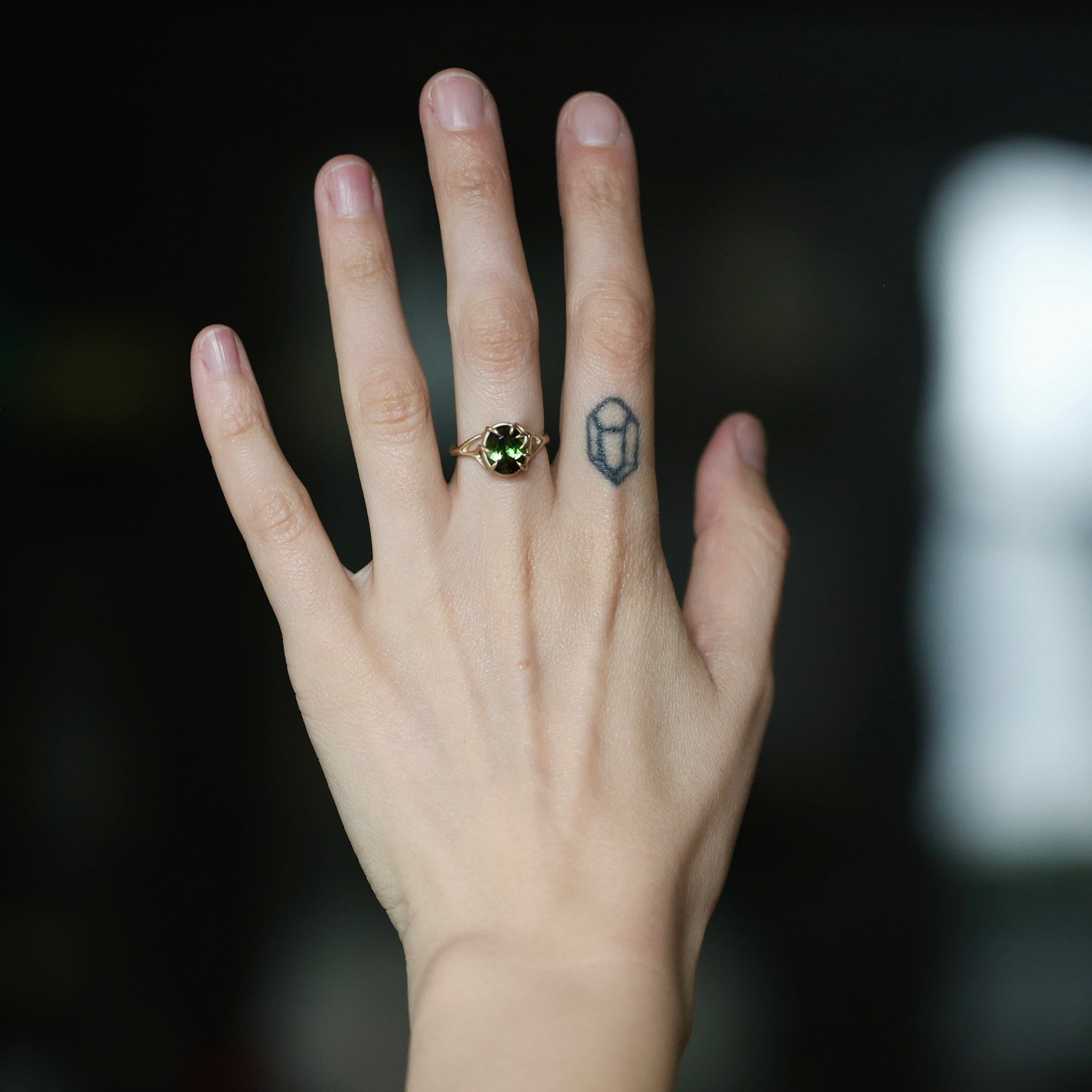 A close-up of a person's hand wearing a gold ring with a green gemstone, set against a dark, blurred background.
