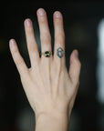 A close-up of a person's hand wearing a gold ring with a green gemstone, set against a dark, blurred background.