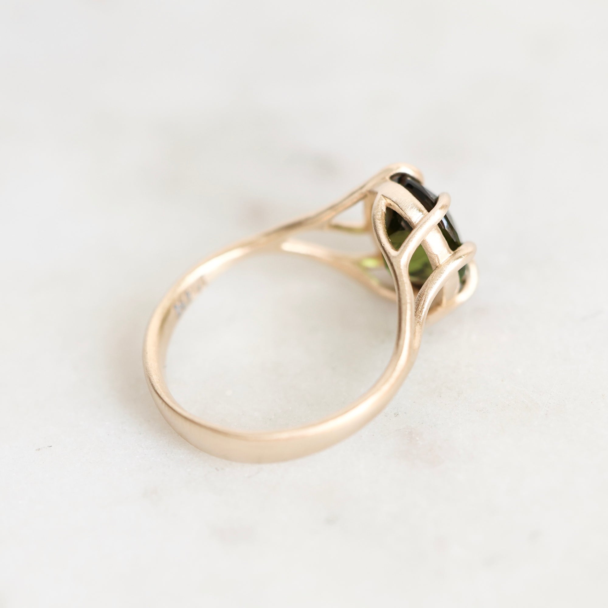 A gold ring with a green gemstone in a split-prong setting, photographed on a light marble surface.