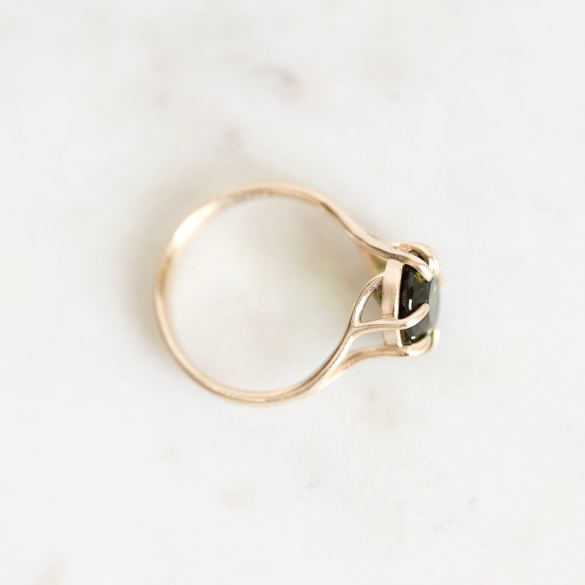 A gold ring with a bezel-set black gemstone, photographed from an angled top-down view on a light marble surface.