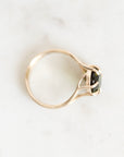 A gold ring with a bezel-set black gemstone, photographed from an angled top-down view on a light marble surface.