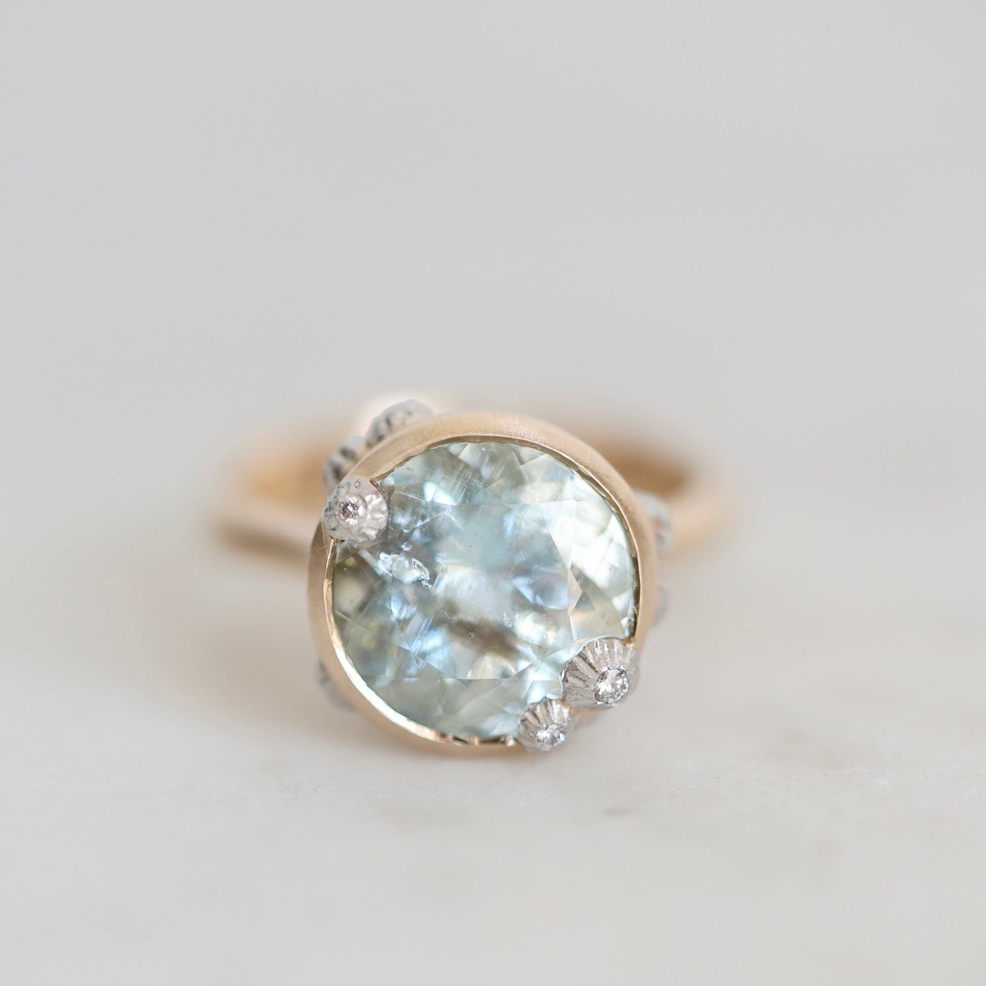 A round bezel-set ring featuring a light blue aquamarine gemstone in a gold-toned setting, photographed on a light gray background.