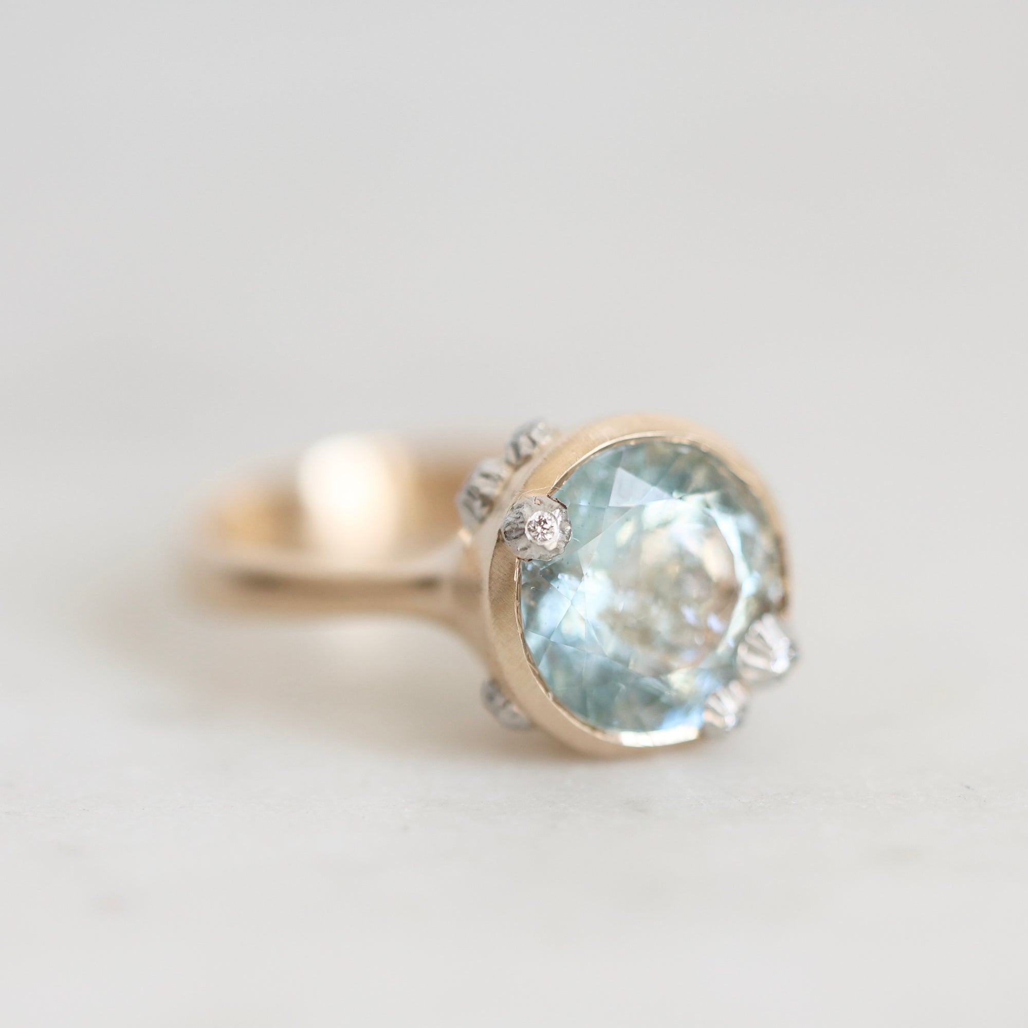 A close-up shot of a gold ring featuring a large oval light blue gemstone, accented with small clear stones on the shoulders, set against a soft, light gray background.