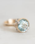 A close-up shot of a gold ring featuring a large oval light blue gemstone, accented with small clear stones on the shoulders, set against a soft, light gray background.