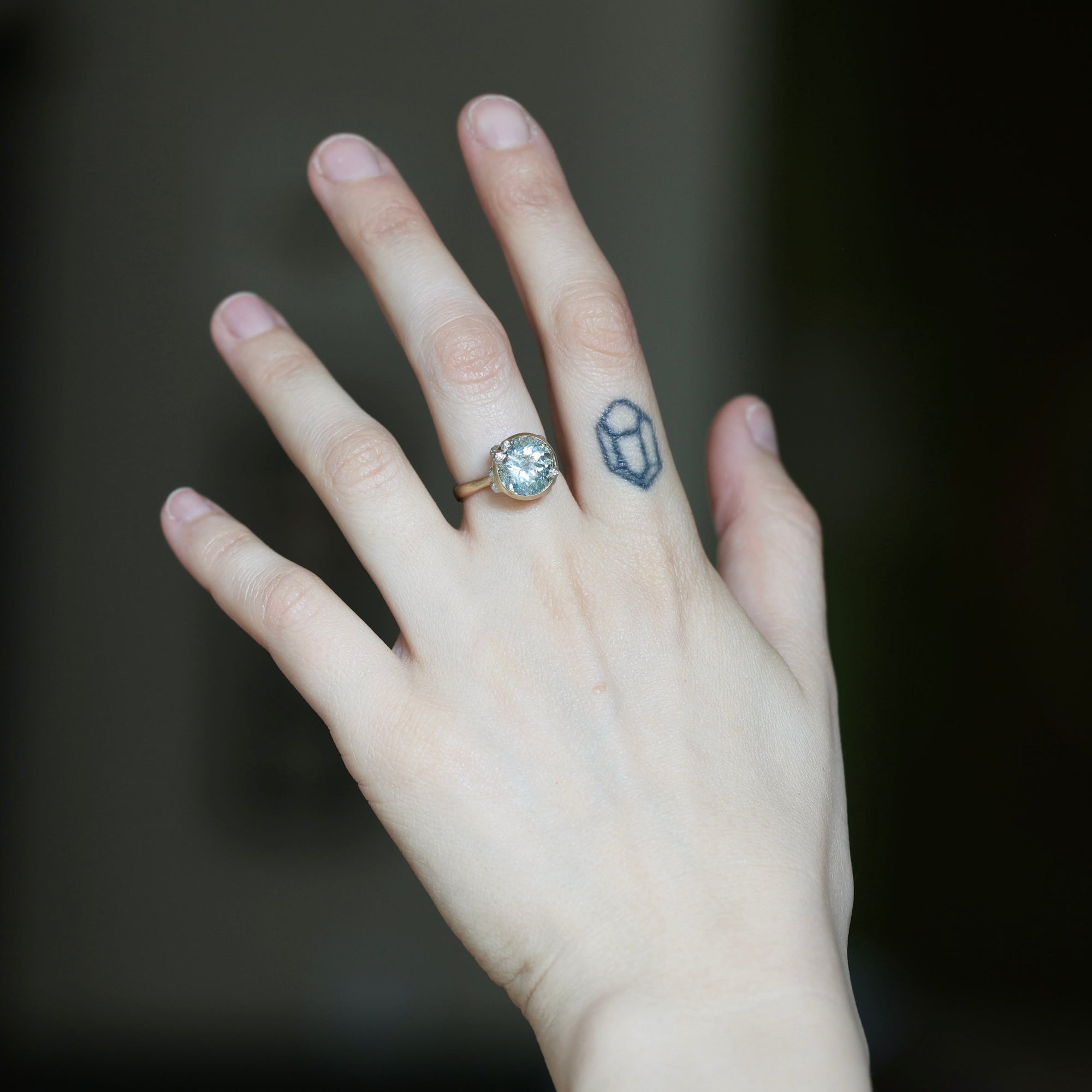 A close-up of a hand wearing a gold ring with a clear round blue with a small blue geometric tattoo on the middle finger against a dark blurred background.
