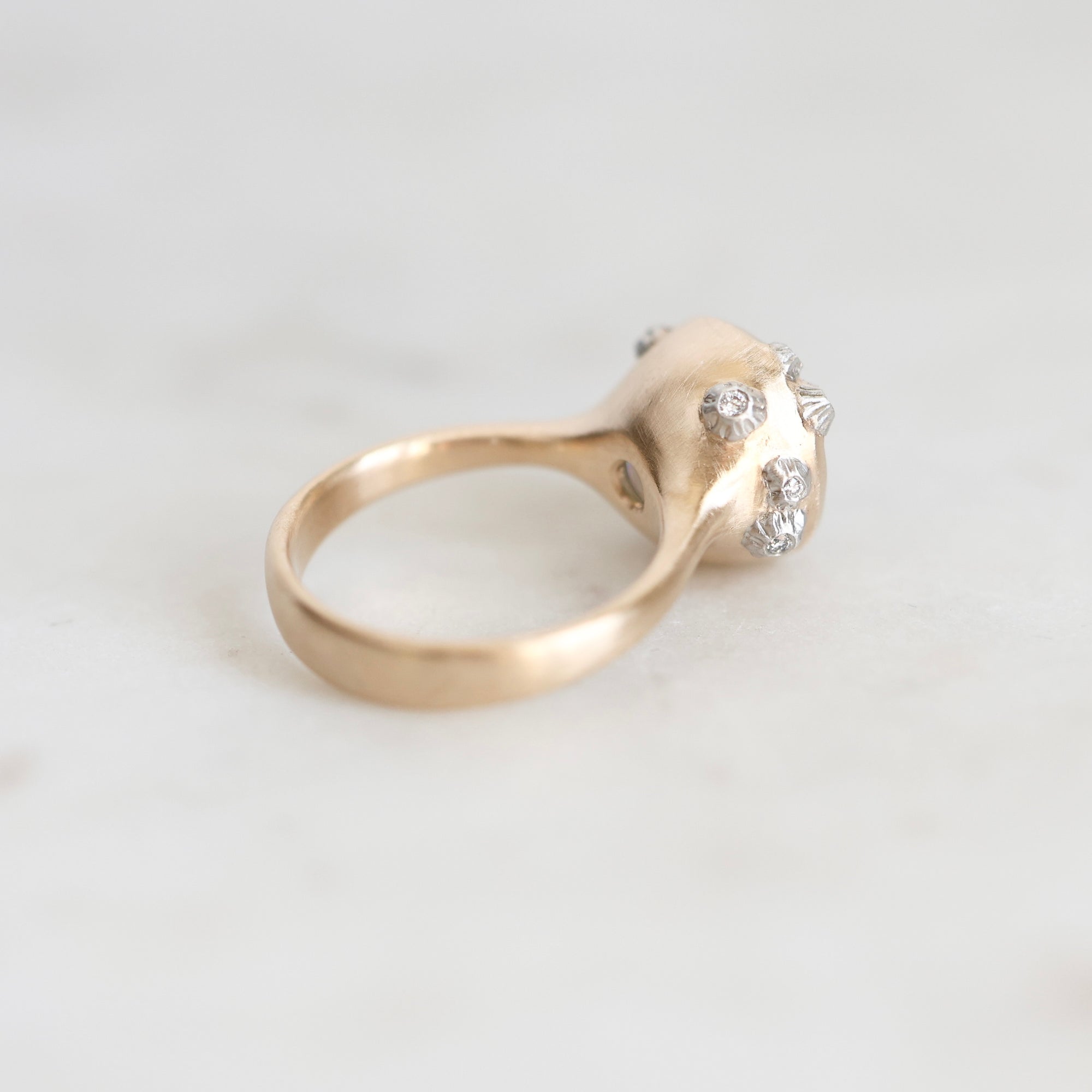 A gold ring with a sculptural, organic-shaped top set with small clear gemstones, resting on a light gray marble surface.