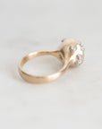 A gold ring with a sculptural, organic-shaped top set with small clear gemstones, resting on a light gray marble surface.