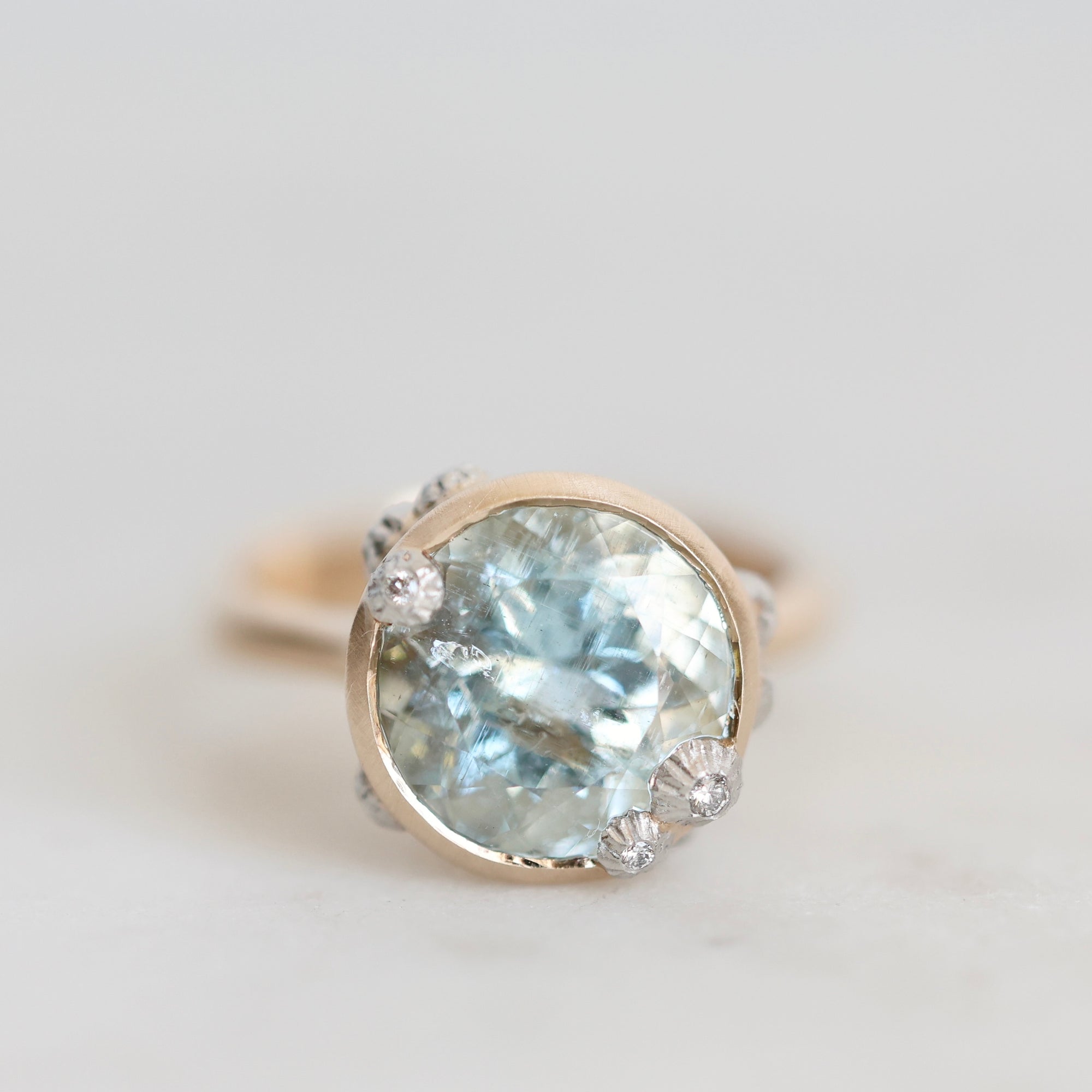 A close-up shot of a vintage-style ring featuring a large round light blue gemstone in a gold bezel setting, accented, set against a soft light gray background.