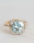 A close-up shot of a vintage-style ring featuring a large round light blue gemstone in a gold bezel setting, accented, set against a soft light gray background.