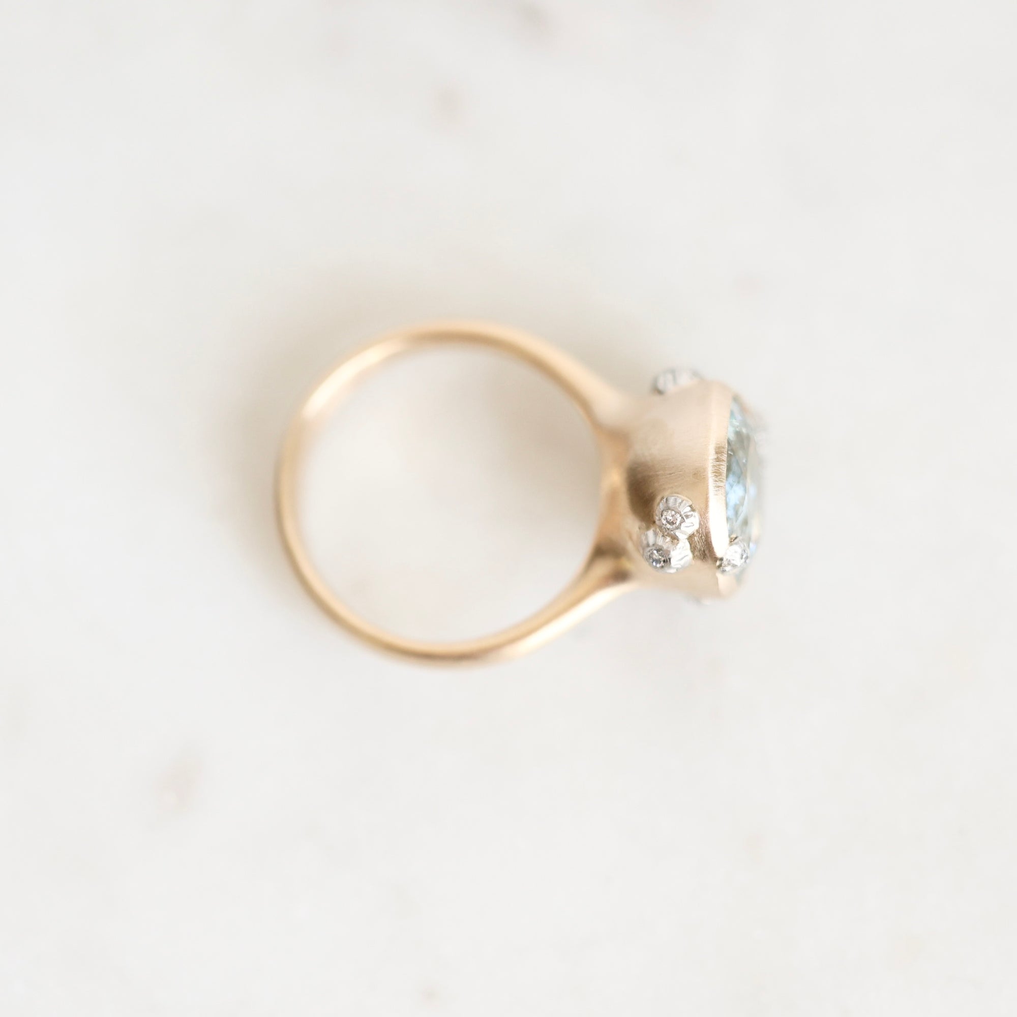 A gold ring with a large oval light blue gemstone and small clear accent stones, photographed on a light marble-like surface.