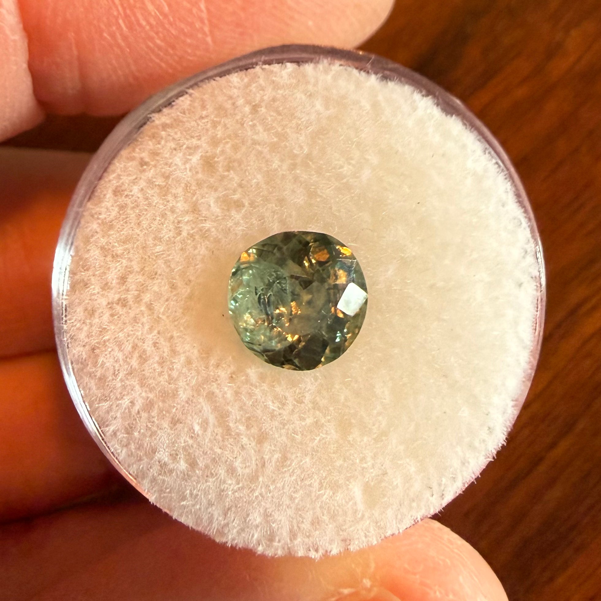 Round greenish gemstone on a white cushion held between fingers with a wooden surface in the background