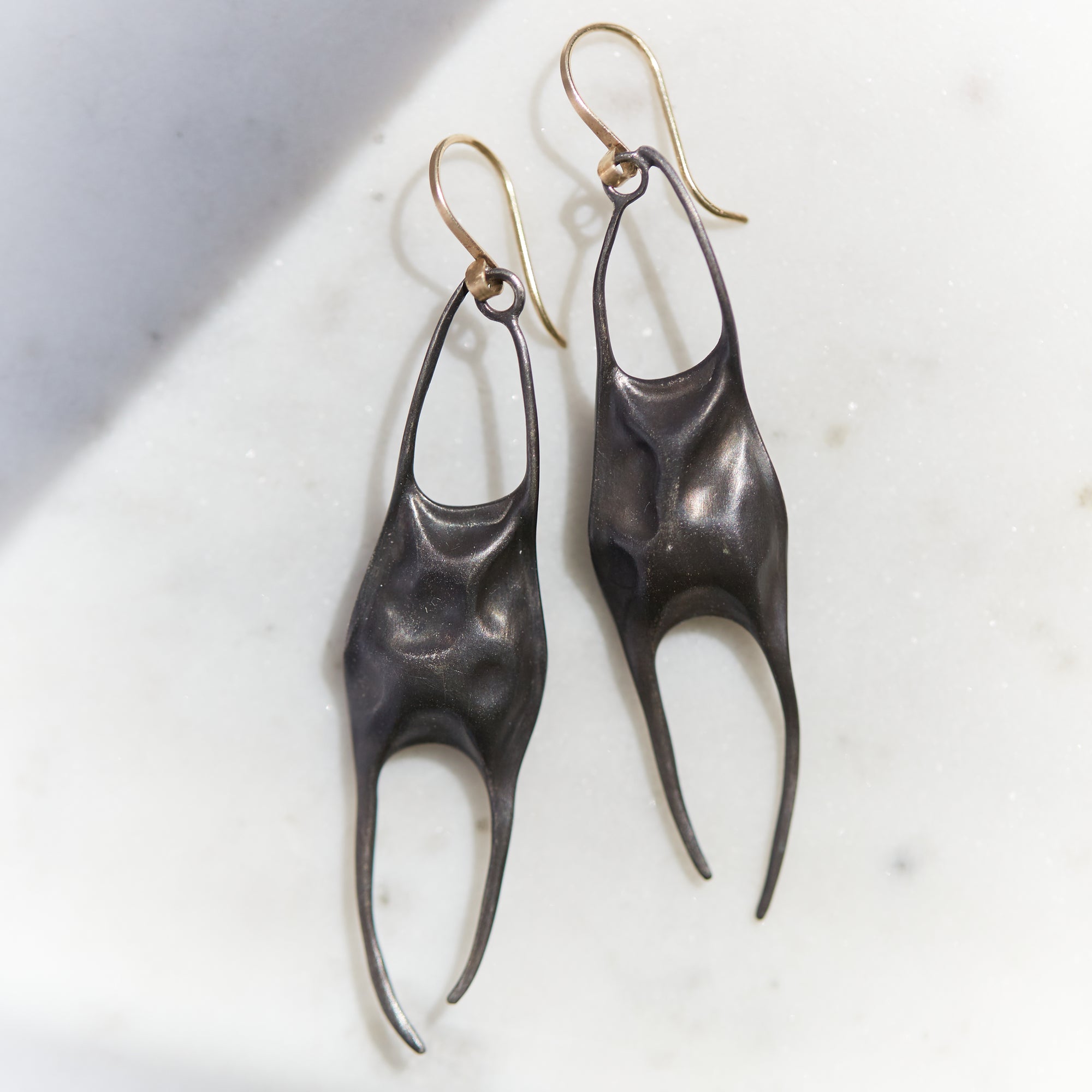 A pair of sculptural dangle earrings with dark oxidized metal bodies and gold-tone hook ear wires, resting on a light marble surface.