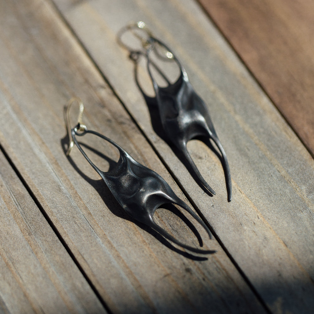 A pair of sculptural black metal earrings with a matte finish, displayed on weathered wooden planks in bright natural sunlight.