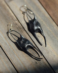 A pair of sculptural black metal earrings with a matte finish, displayed on weathered wooden planks in bright natural sunlight.