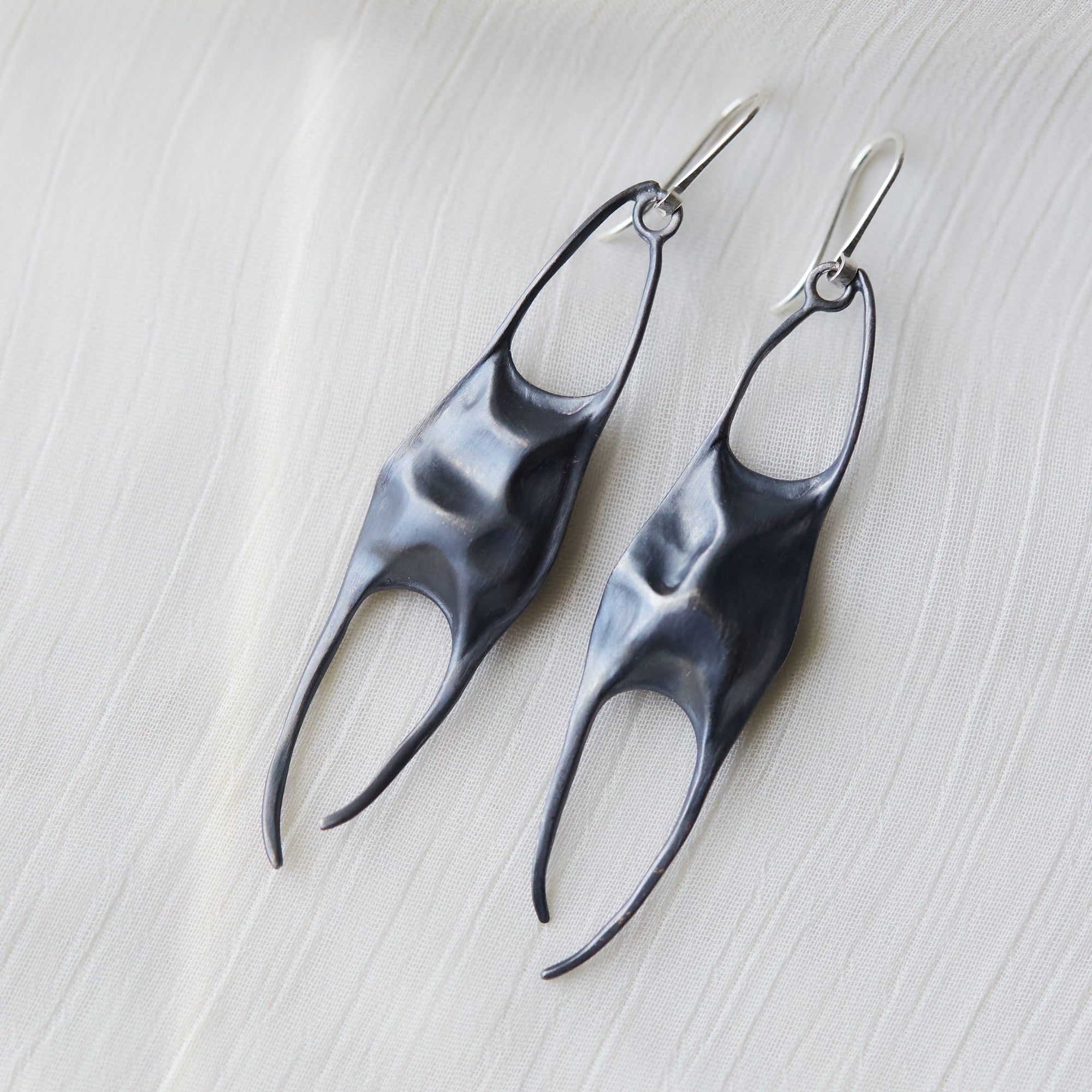A pair of sculptural, organic-shaped drop earrings in a dark metallic finish, displayed on a textured white fabric background.