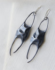 A pair of sculptural, organic-shaped drop earrings in a dark metallic finish, displayed on a textured white fabric background.