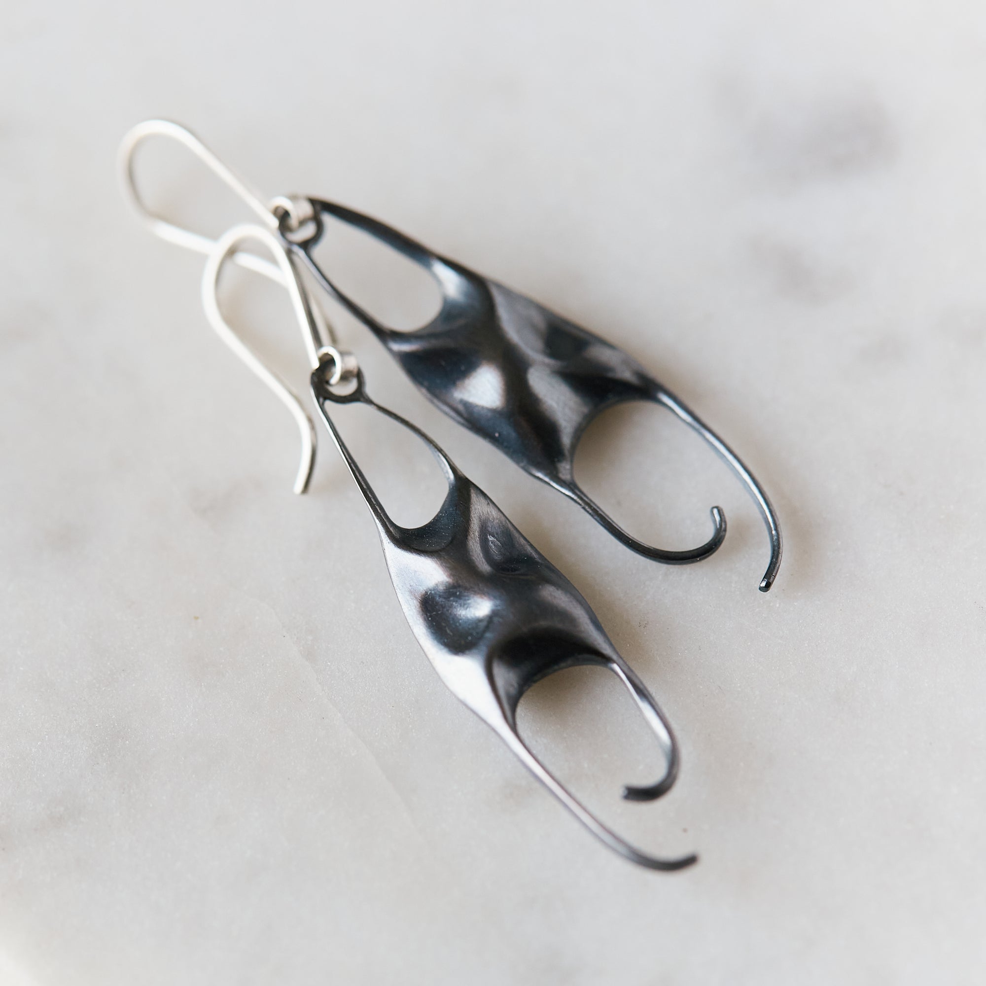 A pair of sculptural dangle earrings with organic, flowing shapes in bright silver and dark oxidized black, resting on a light marble surface.