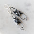 A pair of sculptural dangle earrings with organic, flowing shapes in bright silver and dark oxidized black, resting on a light marble surface.