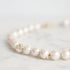 A close-up of a pearl strand necklace resting on a light marble surface. The pearls are white with subtle luster and photographed against a soft, out-of-focus light background
