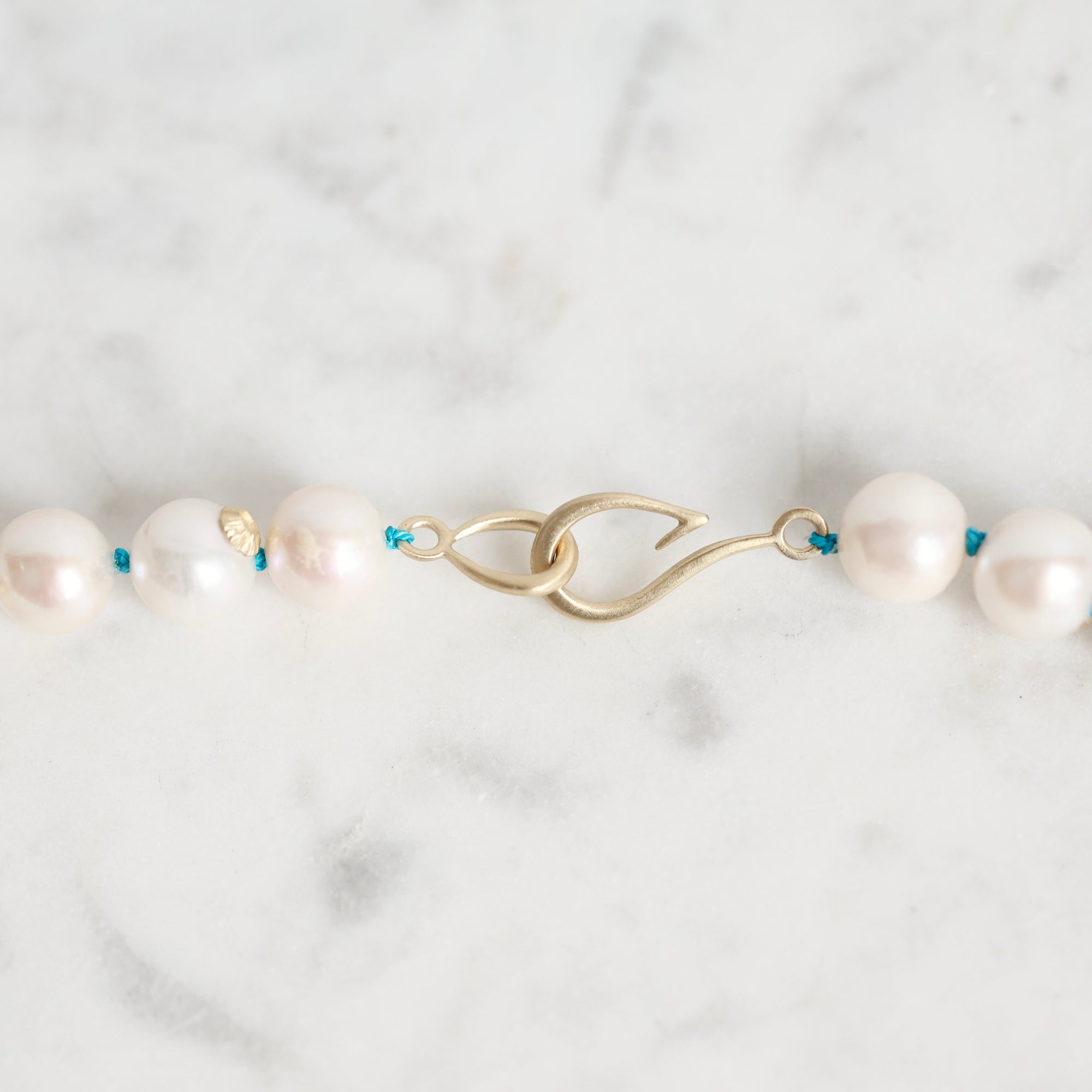 A close-up of a pearl necklace with white round pearls threaded on a gold-toned chain, featuring a gold clasp and small blue accents, laid on a light marble surface.
