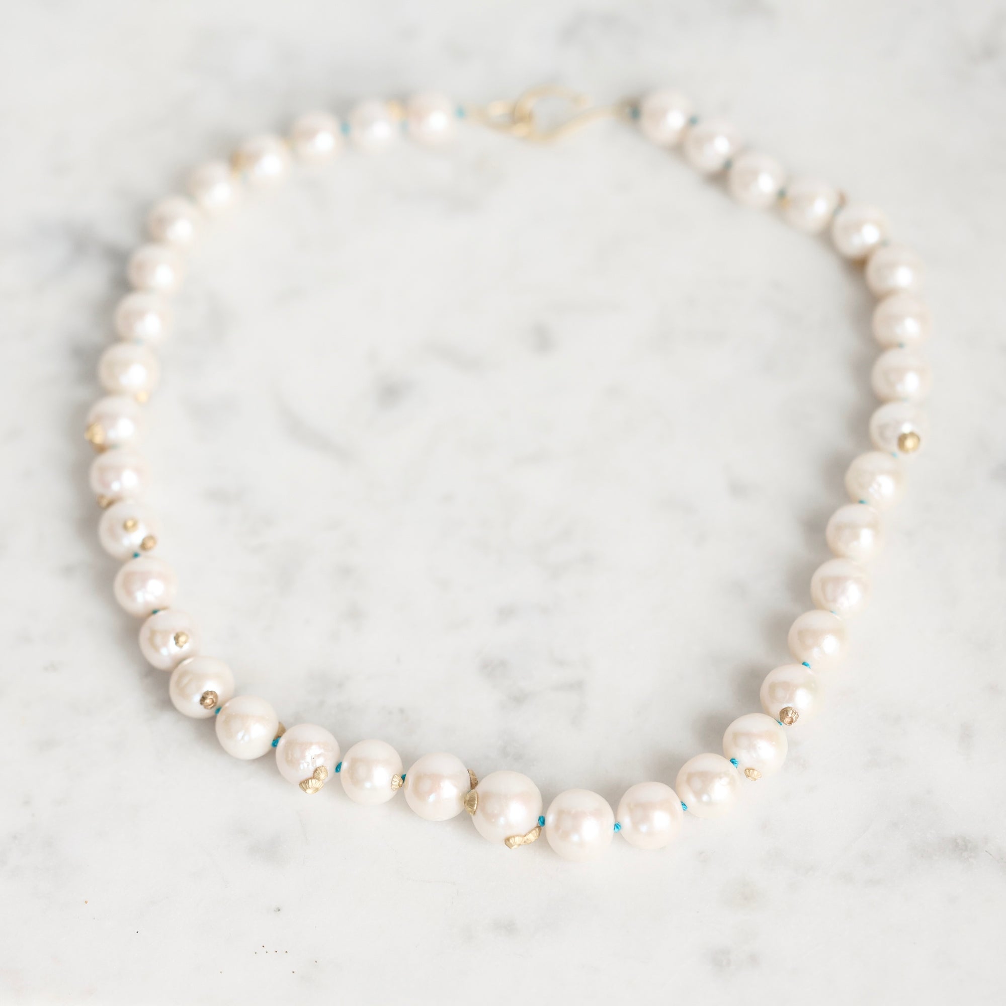 A strand pearl necklace with small gold barnacles, arranged in a circle on a white marble surface.