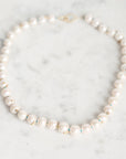 A strand pearl necklace with small gold barnacles, arranged in a circle on a white marble surface.