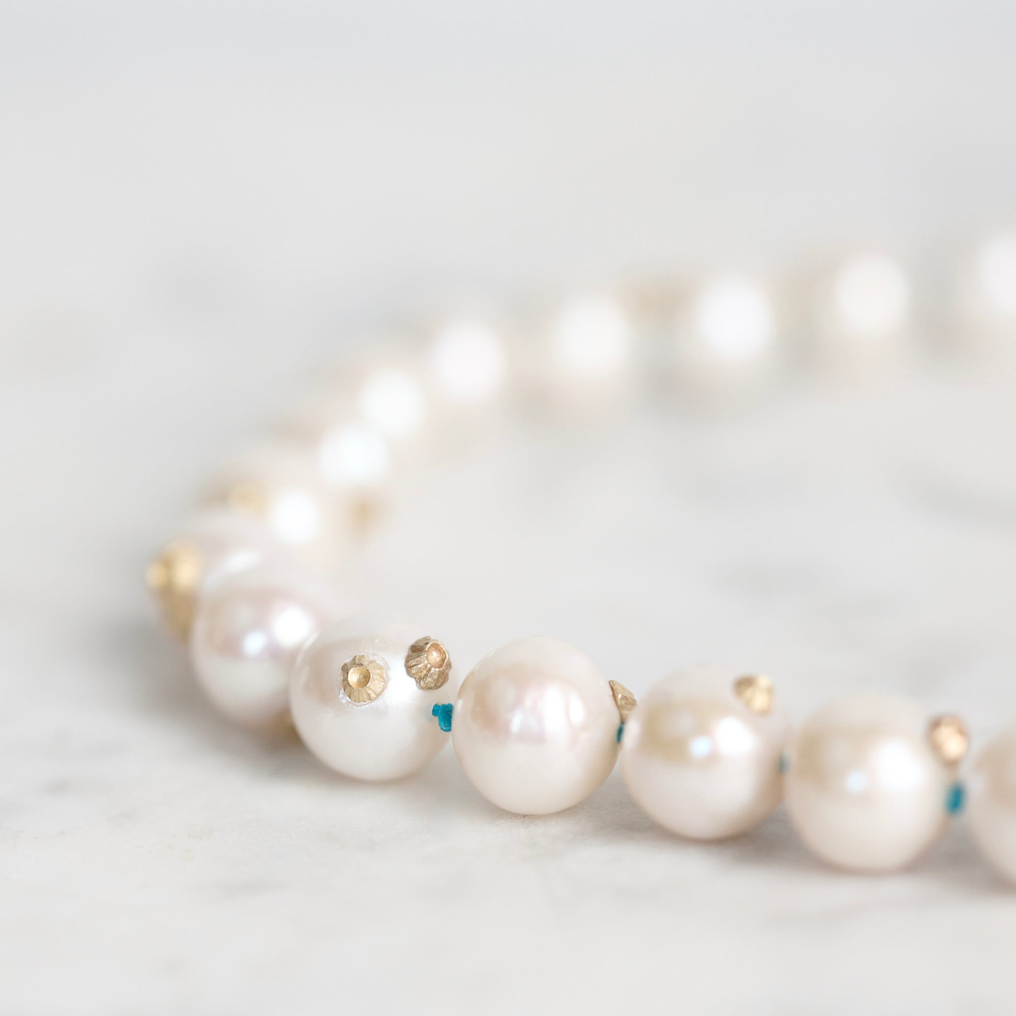 A close-up of a pearl strand necklace with small gold and blue bead accents, resting on a light marble surface.