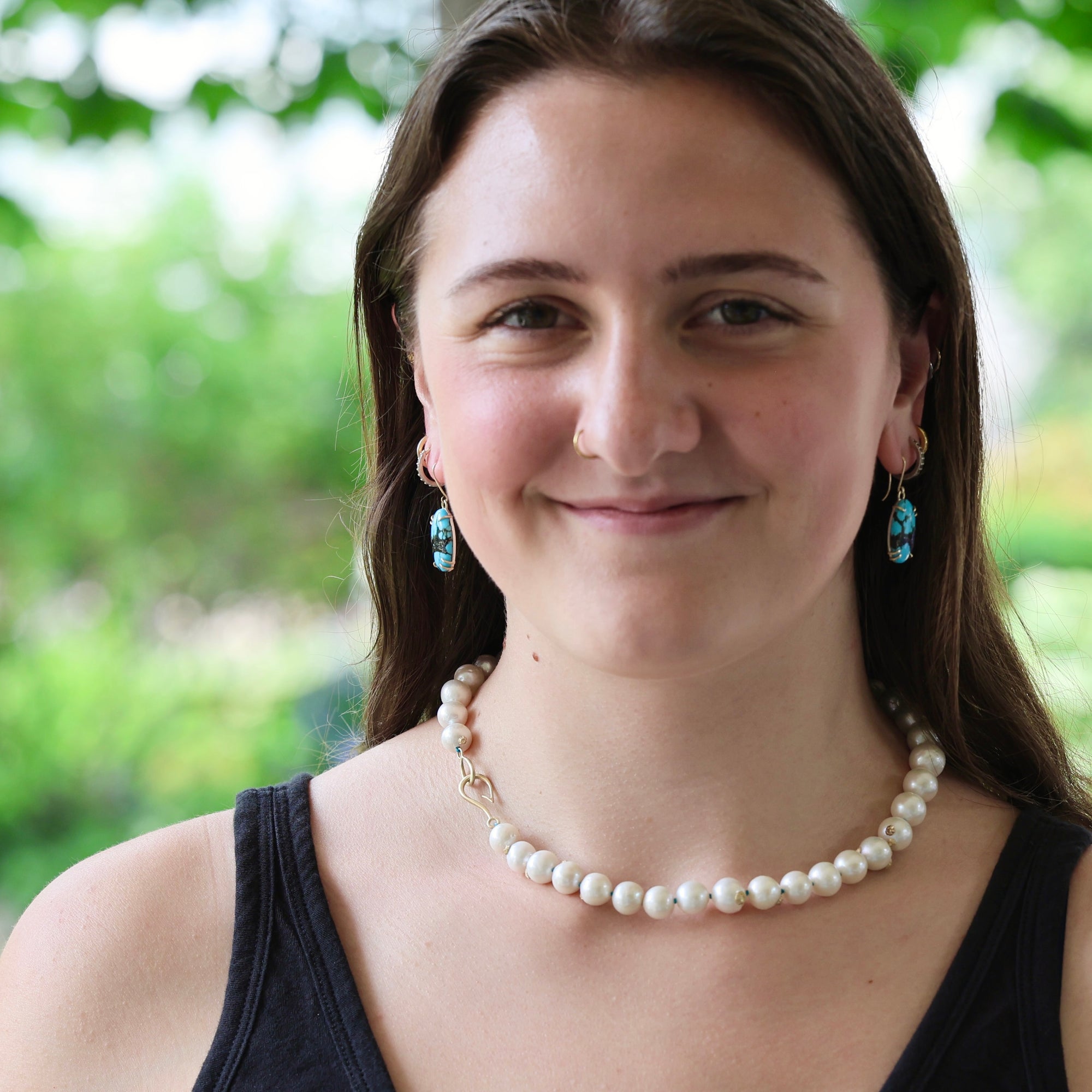 A woman with long brown hair smiling, wearing a white pearl necklace and matching drop earrings with blue stones, set against a soft green outdoor background.