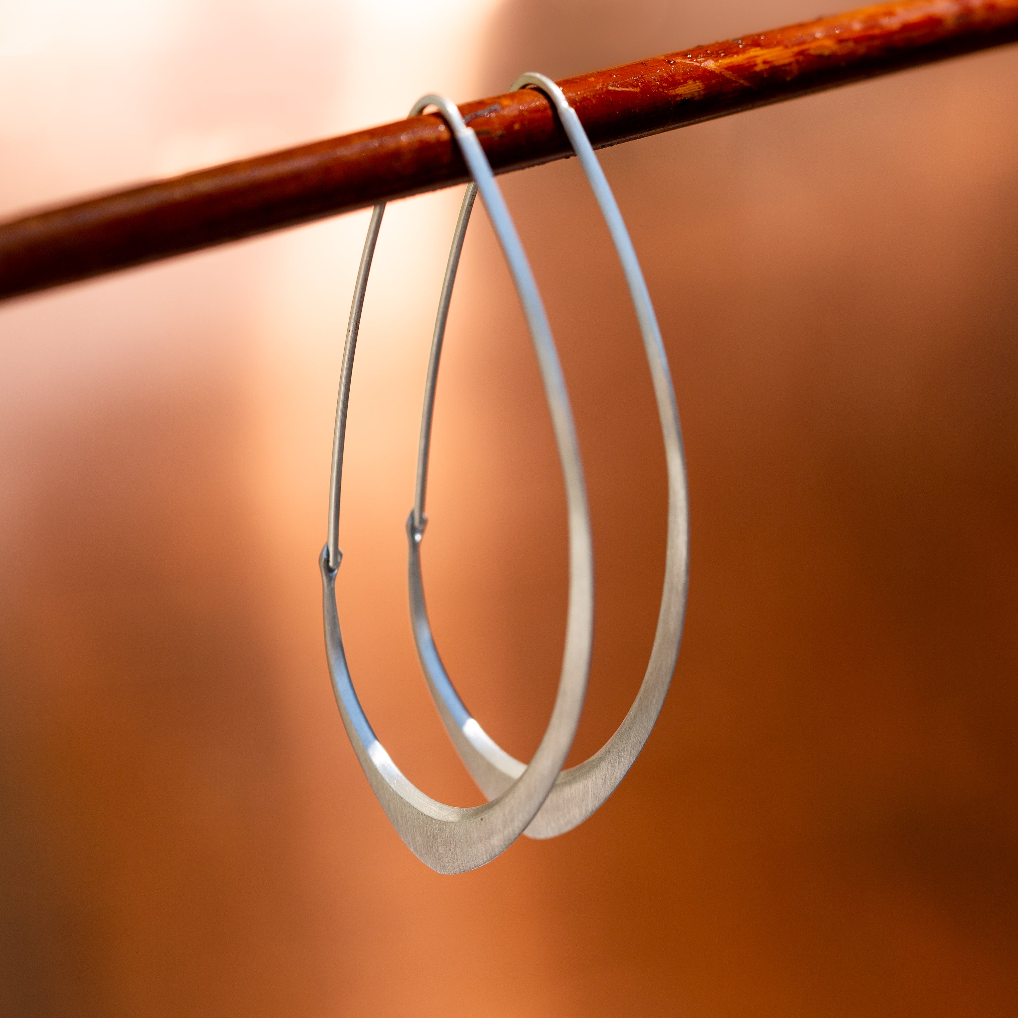 Large Facet Hoops in silver hanging from a wood stick, set against an orange background- Hannah Blount Jewelry